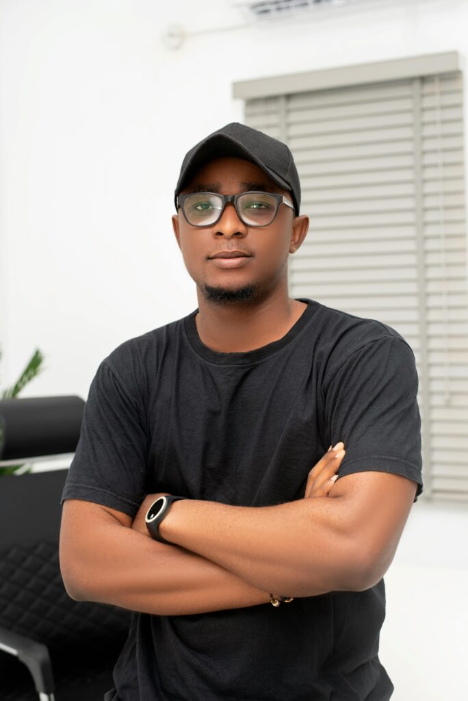 Portrait of a confident man indoors wearing a black hat and glasses with arms crossed.